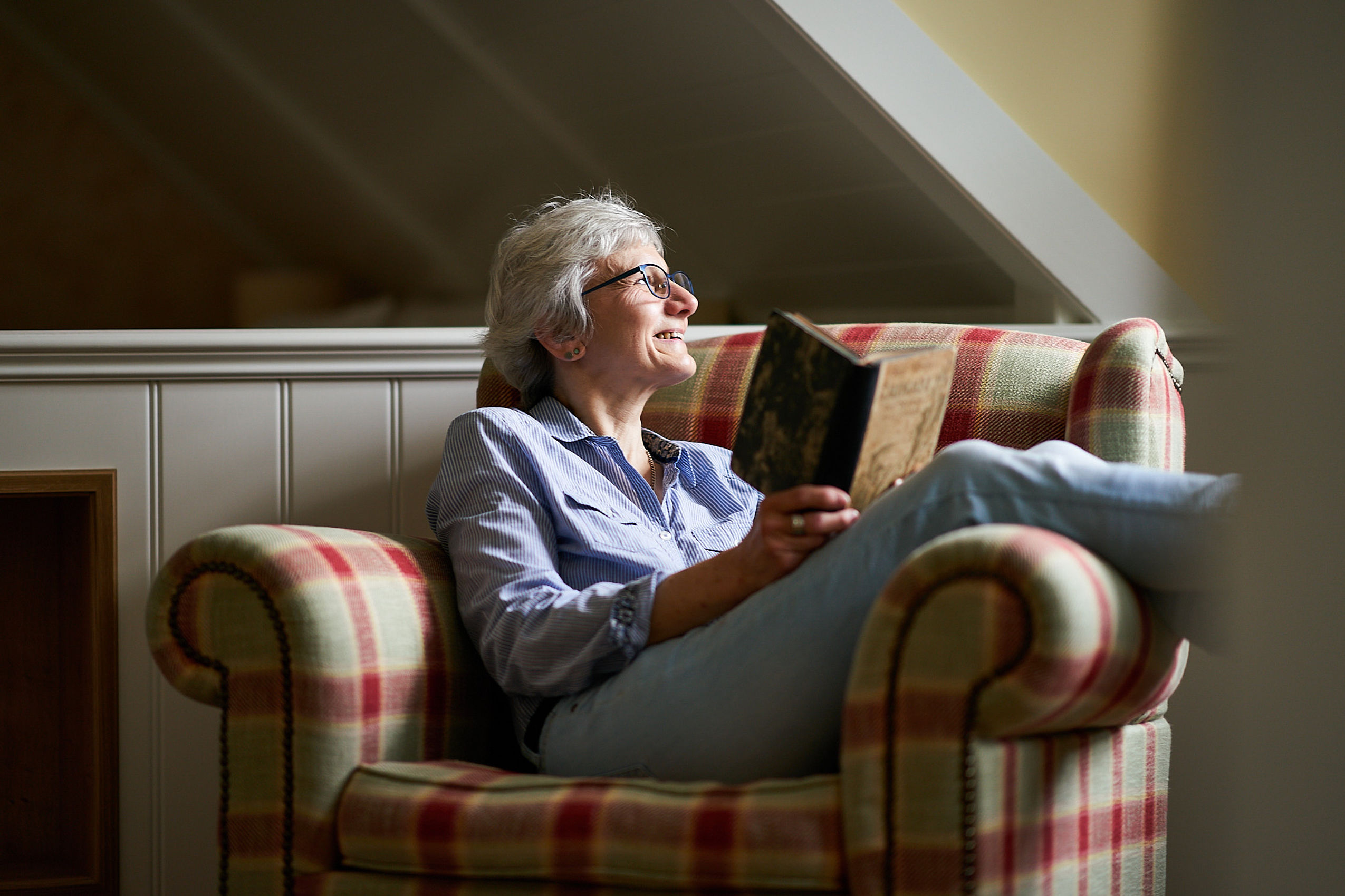 Eine ältere Frau liest auf einem karierten Stuhl ein Buch und schaut lächelnd durch das Fenster