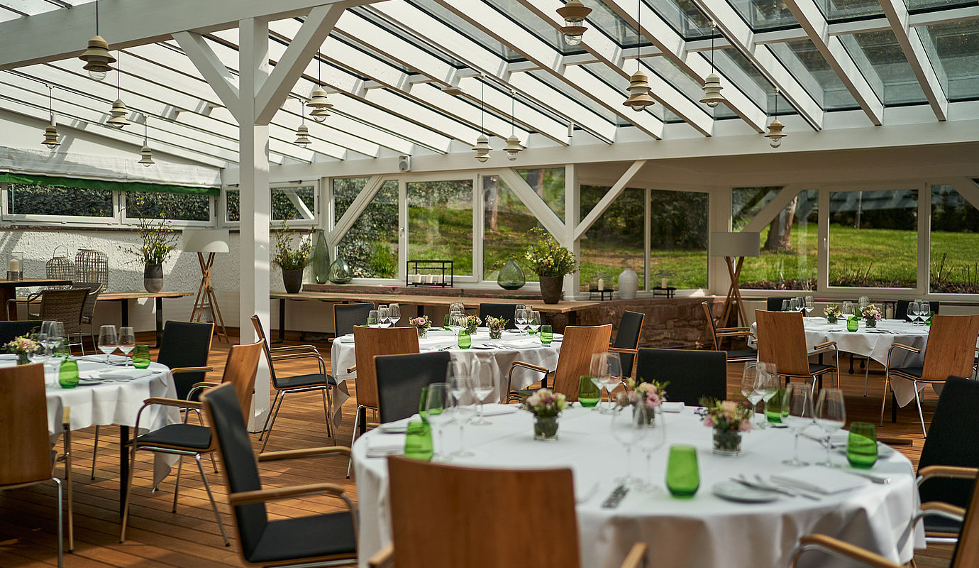 Stühle und runde Tische mit weißen Tischdecken und Blumenschmuck für eine Veranstaltung im Raum mit großen Fenstern mit Gartenblick
