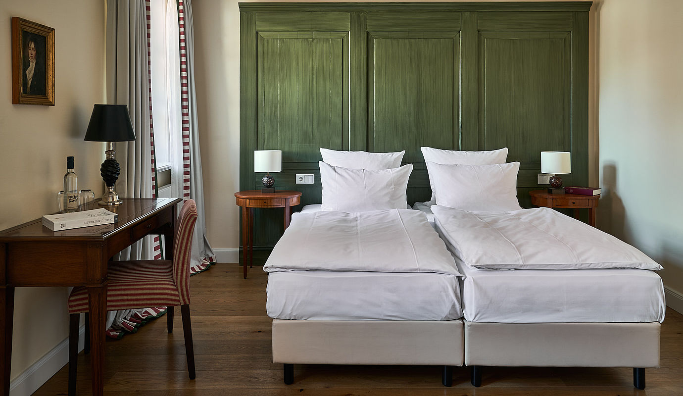 Ein Doppelbett vor einer grünen Wand, ein Holztisch und ein Stuhl neben einem Fenster im Doppelzimmer Cozy