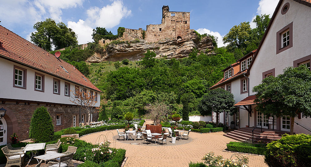Idyllischer Innenhof eines Hotels mit Gartentischen und Blick auf eine mittelalterliche Burgruine.