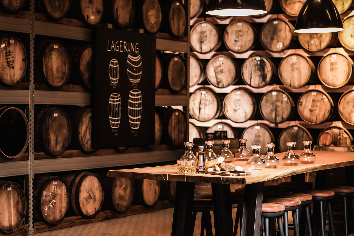 Nahaufnahme eines Tisches mit Flaschen und Gläsern, umgeben von Weinfässern in der Hardenberg Distillery