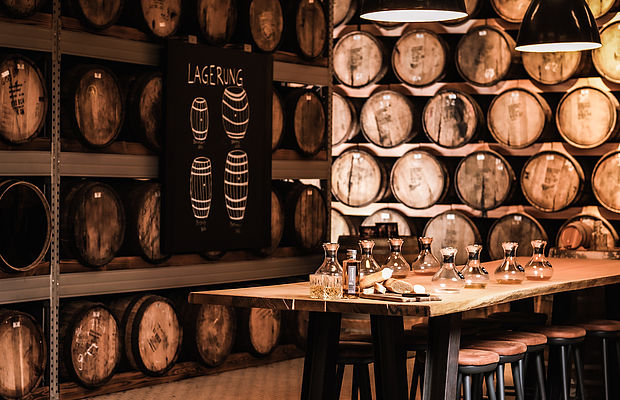 Nahaufnahme eines Tisches mit Flaschen und Gläsern, umgeben von Weinfässern in der Hardenberg Distillery