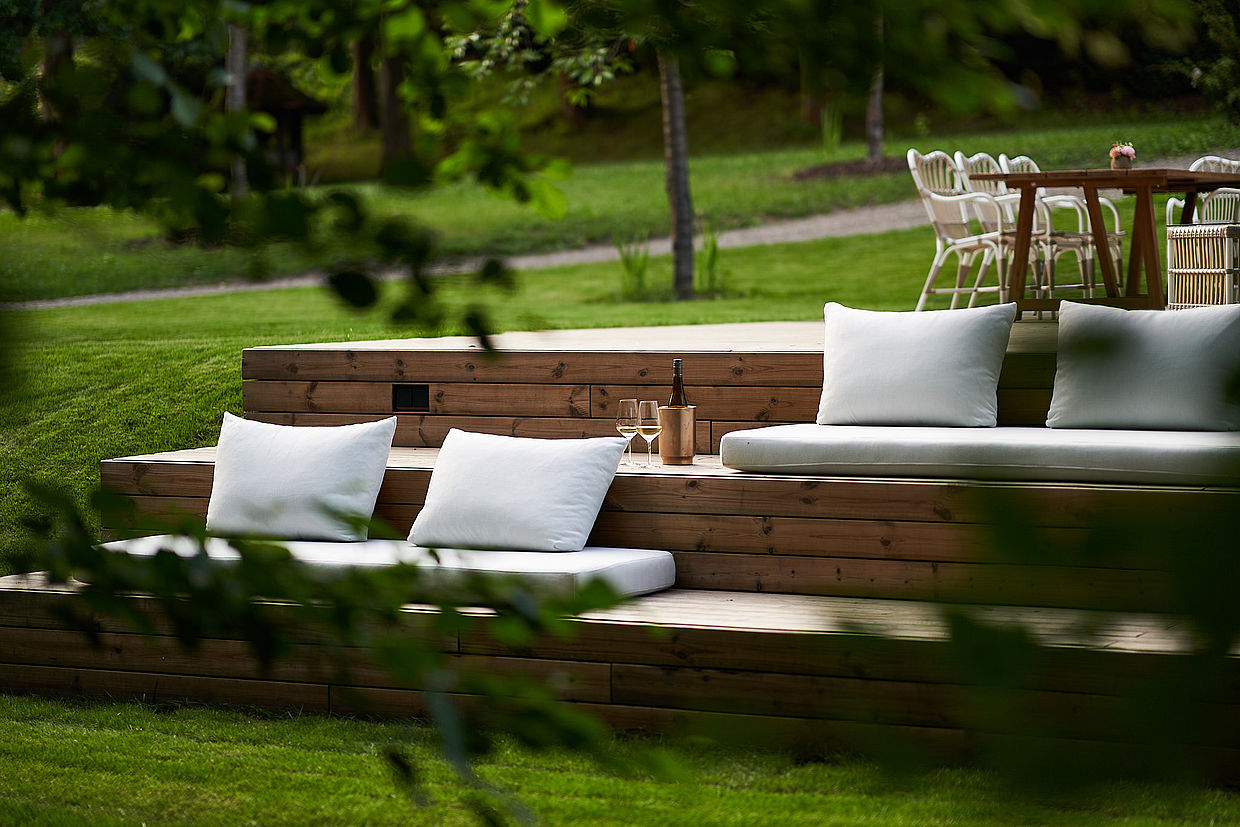 Gemütliche Outdoor-Lounge mit Holzstufen, weißen Kissen und einer Flasche Wein in einem grünen Garten
