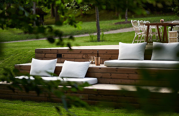 Gemütliche Outdoor-Lounge mit Holzstufen, weißen Kissen und einer Flasche Wein in einem grünen Garten
