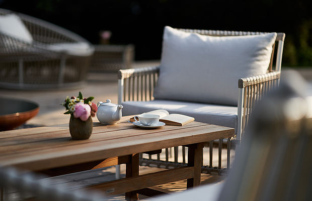 Holztisch auf der Terrasse mit Teekanne, Tasse, Buch und Blumenvase, im Hintergrund ein Sofa